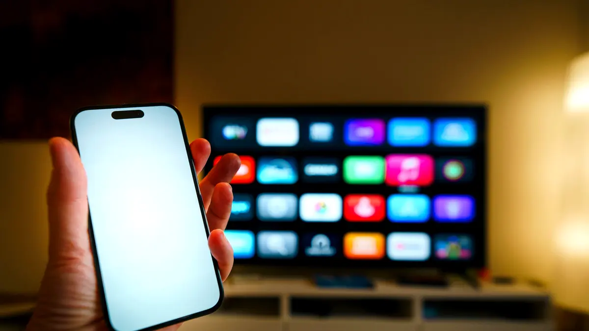A phone held in front of a smart TV showing a grid of streaming apps.