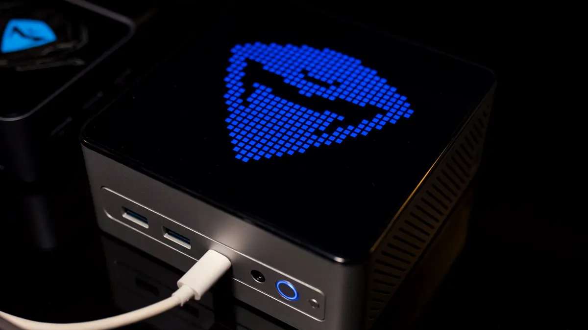 A mini PC on a desk with its status display lit, dwarfed by the monitor behind it.